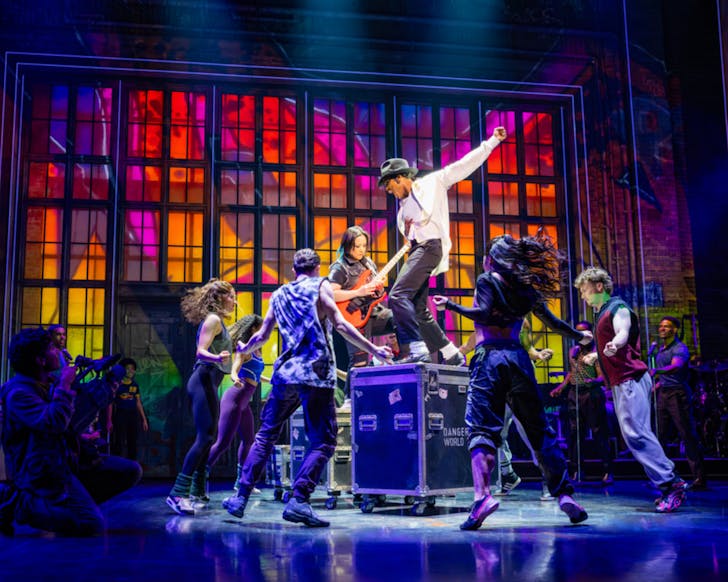 The stage of MJ the musical mid-performance as the actor for Michael Jackson stands elevated on a music equipment box while striking an iconic pose and the dancers are on the floor, lower than him, striking poses and surrounded by neon lighting.