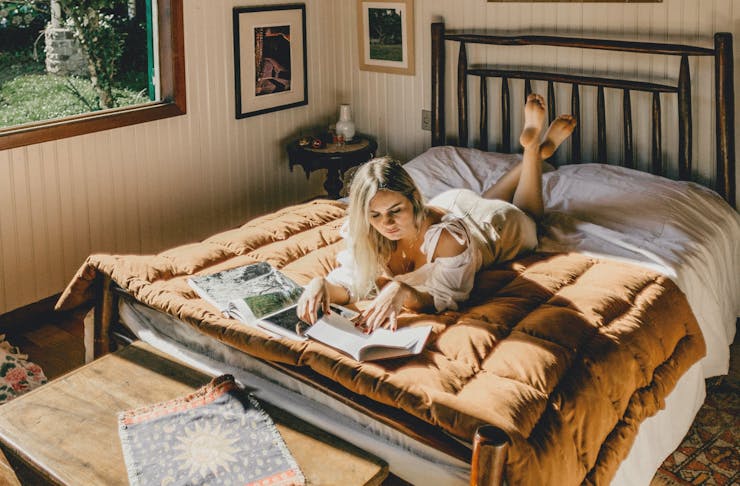 A blonde woman lies on her sun-drenched bed while reading a magazine.