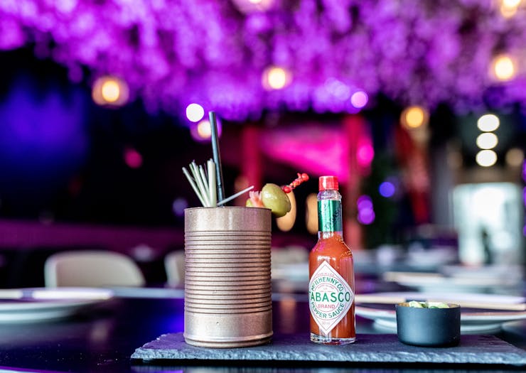 A Bloody Mary style cocktail served in a tin can with a bottle of Tabasco beside it, against a neon-purple backdrop.