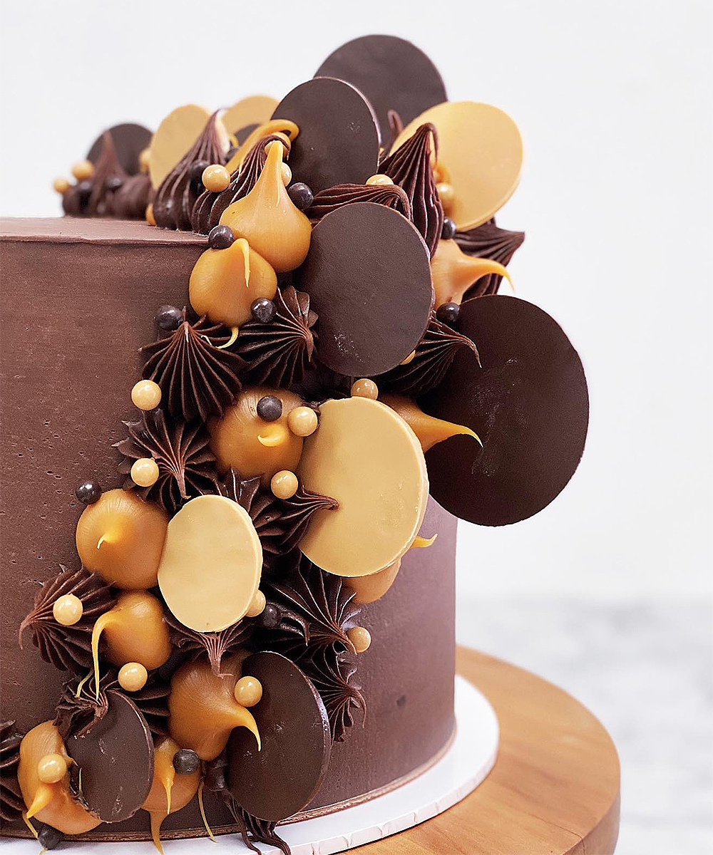 Rich chocolate layered with salted caramel ganache and dark chocolate ganache and decorated with dark chocolate and dulce chocolate disks, dollops and chocolate pearls from Magnolia Kitchen.
