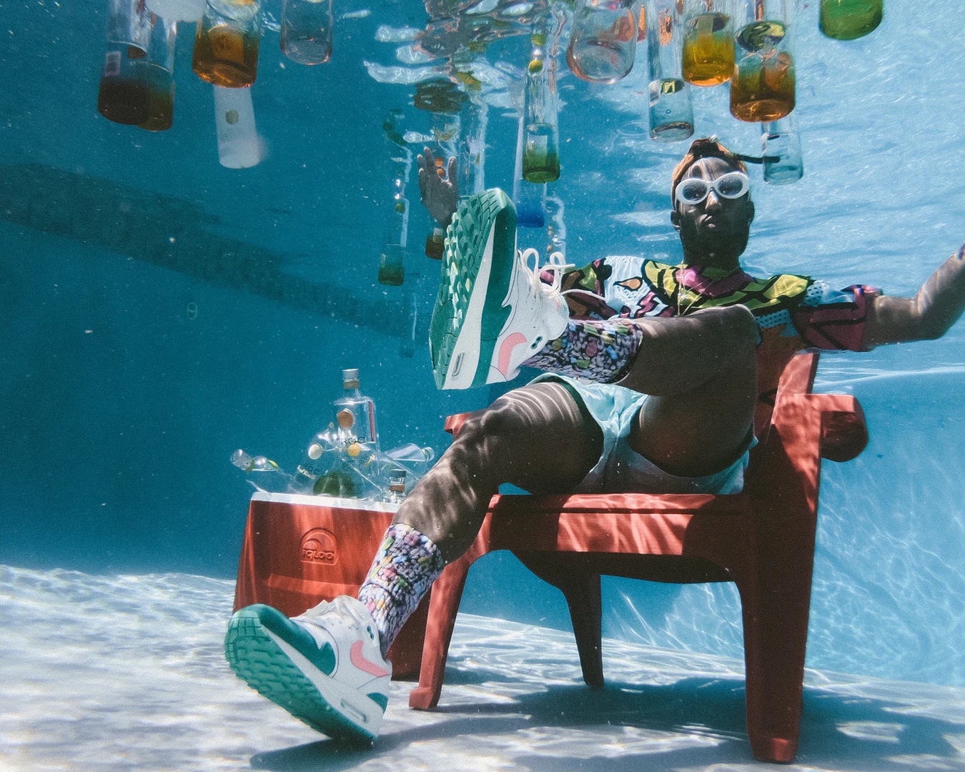 Man underwater sitting on a chair surrounded by floating bottles.
