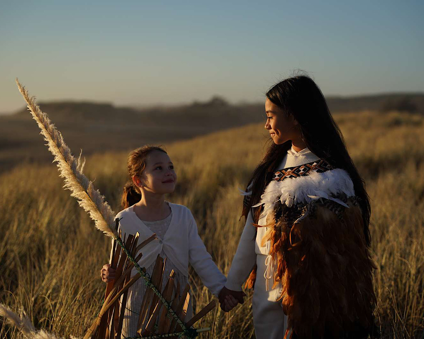 Two young girls, one dressed in traditional Māori clothing are about to fly a kite. 