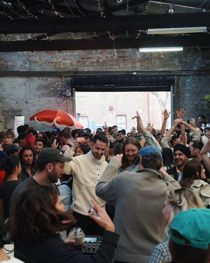 A wide shot of a crowd of people dancing to music played by a DJ in the foreground of the image. The area is indoors with fairy lights decorating the roof and a casual feel, and there is a large open space outside that shows it is daylight. People hold coffees in their hands.