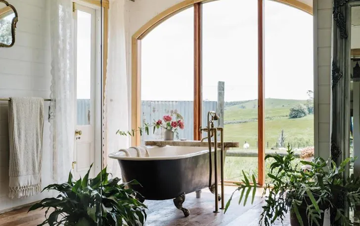 A free-standing bathtub us in the middle of a bathroom looking out through floor-to-ceiling windows to greenery.