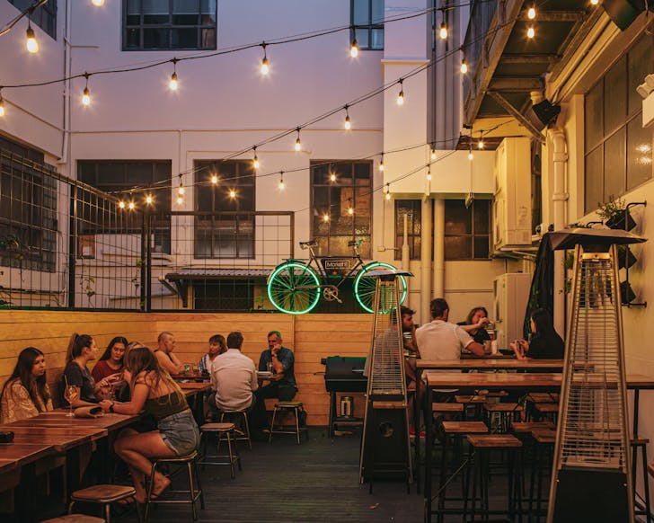 Customers enjoying drinks at the outdoor area of the bar. A cycle with green neon lights hangs on the fence.