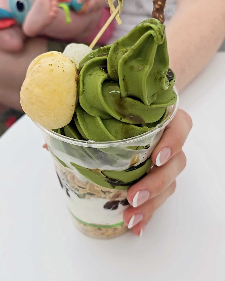 A large plastic cup filled with matcha soft serve and toppings, and a baby's foot and toys in the background. 