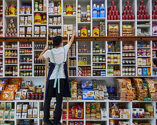 A man reaching on a step to a packet of pasta amongst a wall of different goods.