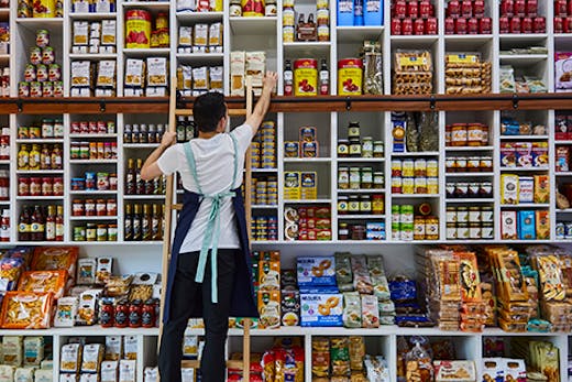 A man reaching on a step to a packet of pasta amongst a wall of different goods.