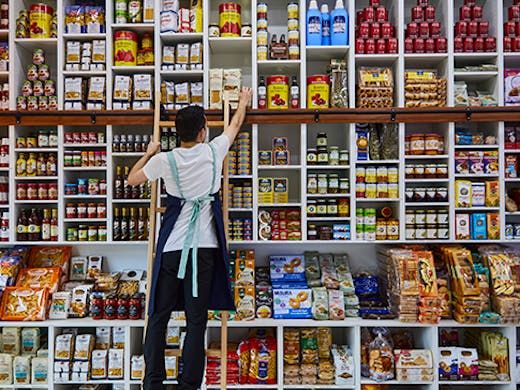 A man reaching on a step to a packet of pasta amongst a wall of different goods.