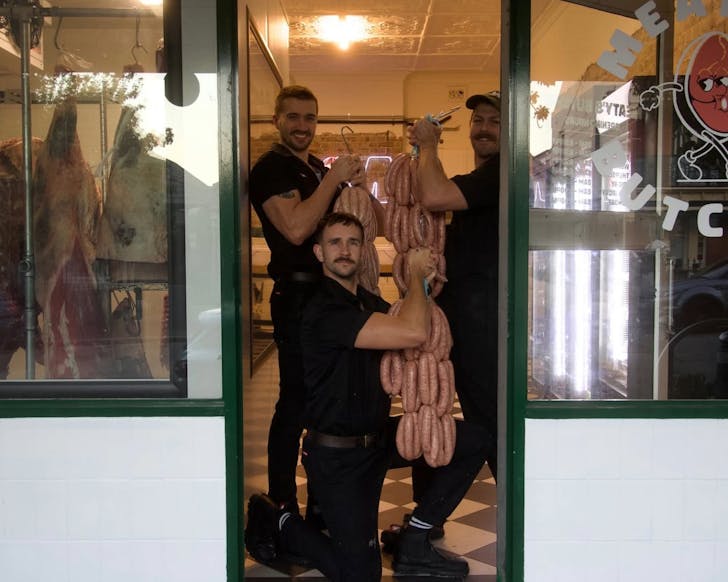 Three men stand in the doorway of Meaty's Butcher. One is kneeling, the other two are standing, and all are holding large portions of butcher meat including sausages. There are all smiling and the poses are slightly comedic.