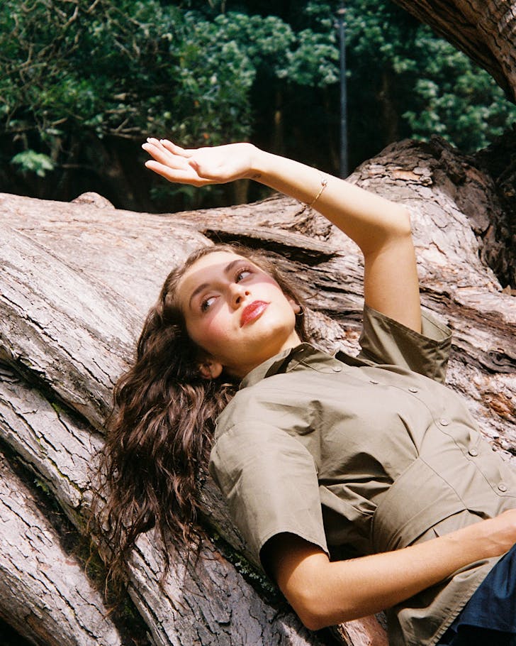 A woman lies up against a tree trunk and uses her hand to block the sun from her eyes.