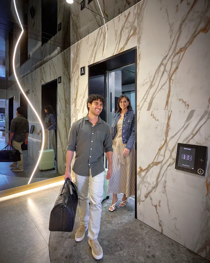 An image of a couple walking out of an elevator in a hotel, holding a luggage bag with big smiles on their faces. Behind them is an elegant lit mirror on the wall that reflects them exiting the elevator, and the other walls are of a nice marble.