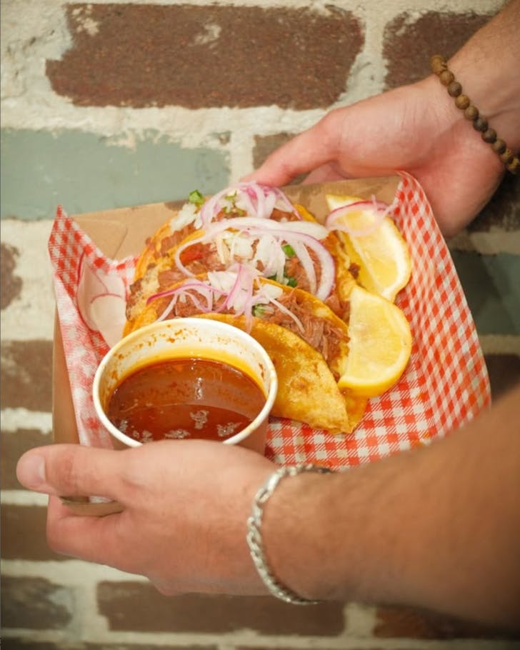 A photo of hands holding a tray of tacos, displayed very nicely and neatly on white and red food paper. 