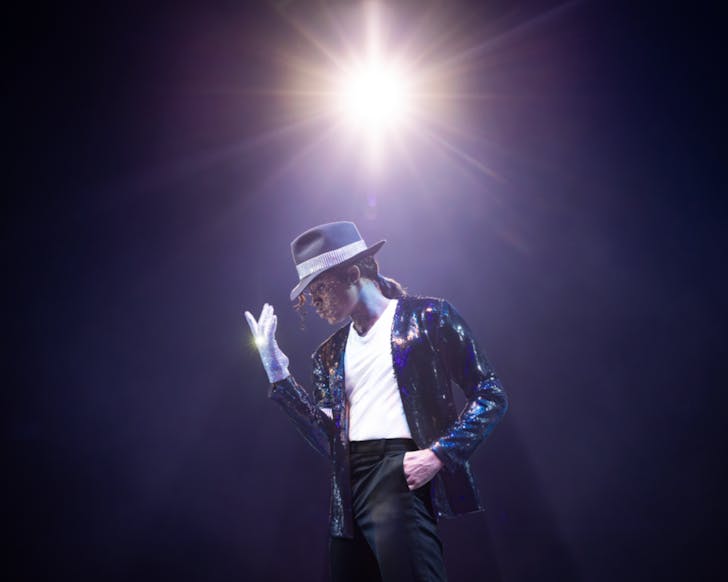 A dark image of a mid-shot of the actor for Michael Jackson in MJ The Musical striking a pose and wearing an iconic sparkly suit. A bright light spotlights him and shines above him in the image. The image is dark and nothing else is visible except the light and MJ.