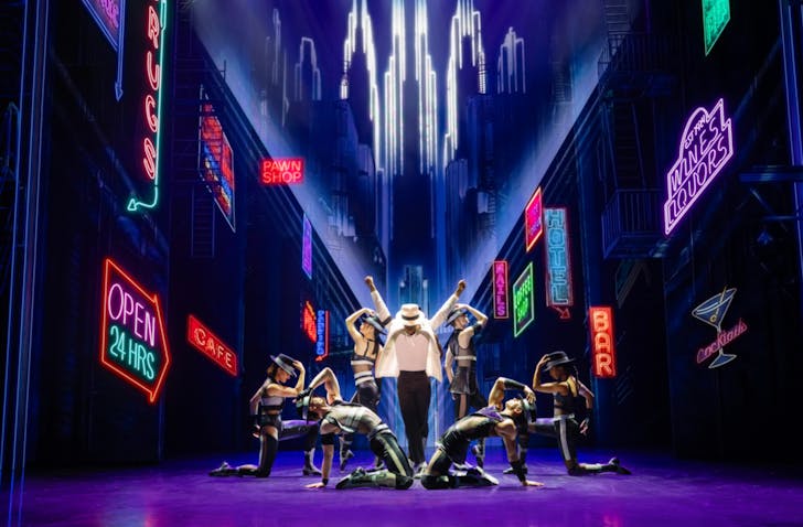 A wide shot of the stage of MJ The Musical mid performance, with the actor of MJ in the middle in an iconic Michael Jackson pose and white suit while neon lights and dancers surround him. He is spotlighted.