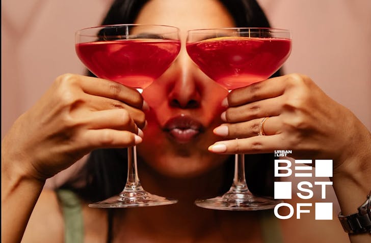 A woman pouts while holding two rose-tinted cocktail glasses in front of her face. 