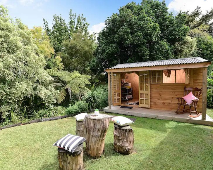 A small wooden cabin adjacent to a gully. A cute table and chair duo made from tree stumps sit in front of this adorable Hamilton airbnb. 