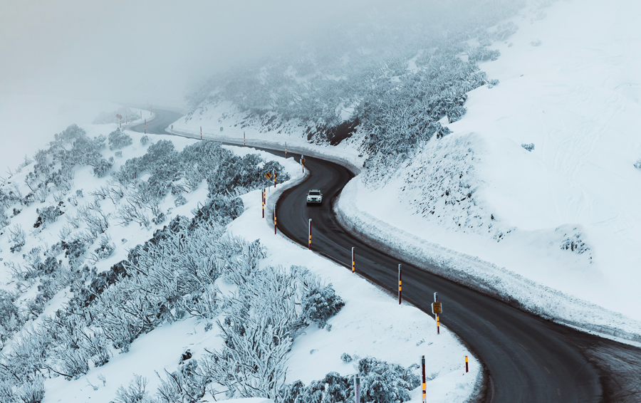A mountain road with snow and a car driving down. 