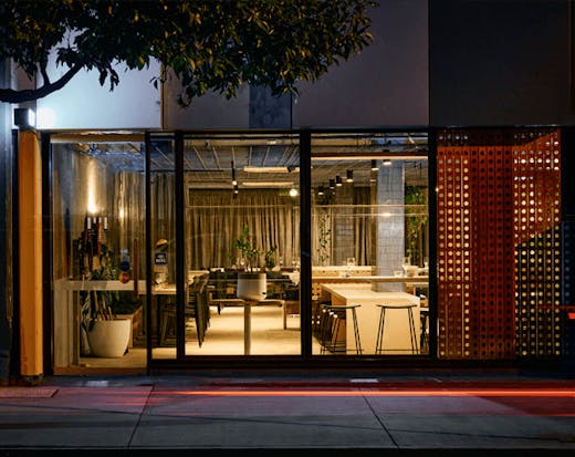 The shopfront of a restaurant with lights on and wooden furniture inside.