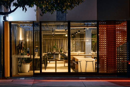 The shopfront of a restaurant with lights on and wooden furniture inside.