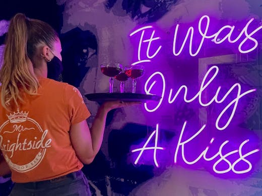 A waitress in an orange Mr Brightside t-shirt holding drinks in front of a purple neon sign that says 'it was only a kiss'