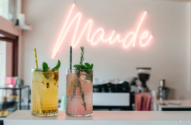 two coloured drinks on a bench in front of a neon sign saying 'maude'