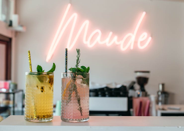 two coloured drinks on a bench in front of a neon sign saying 'maude'