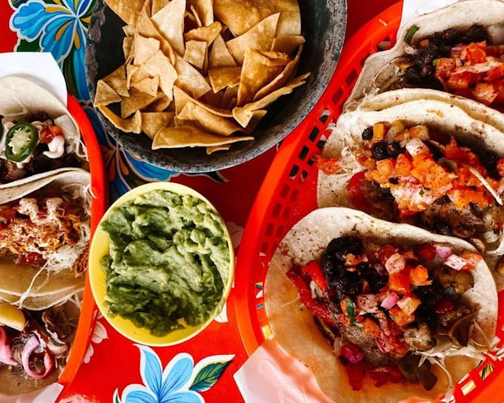 An above view of many trays of Mexican food against a very colourful floral tablecloth.