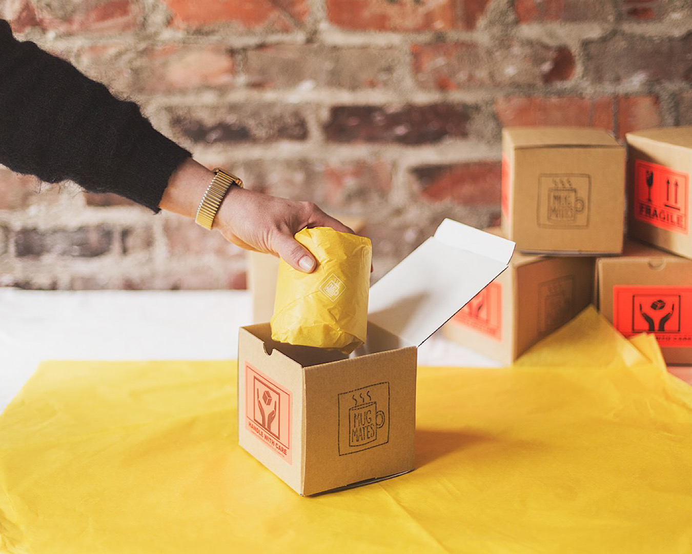 A hand reaches into a cardboard box lifting out a mystery mug wrapped in yellow tissue paper. 