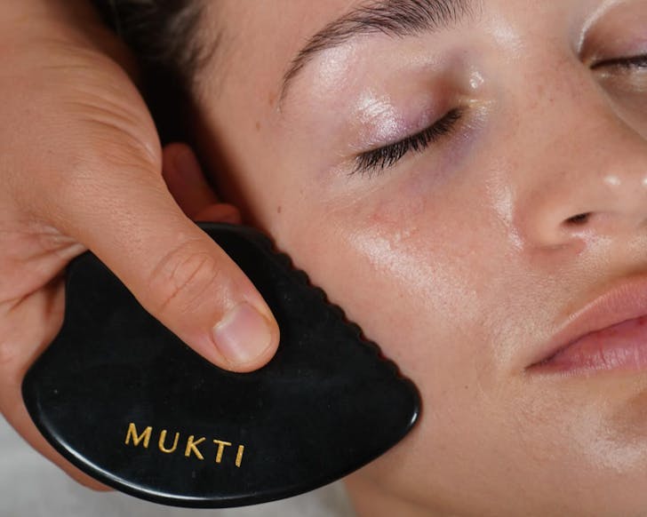 Close up picture of woman's face with hand running black gua sha along her cheek. The gua sha has MUKTI written on it in gold.