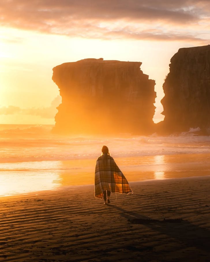 A person wrapped in a blanket walks Muriwai Beach at sunset. 