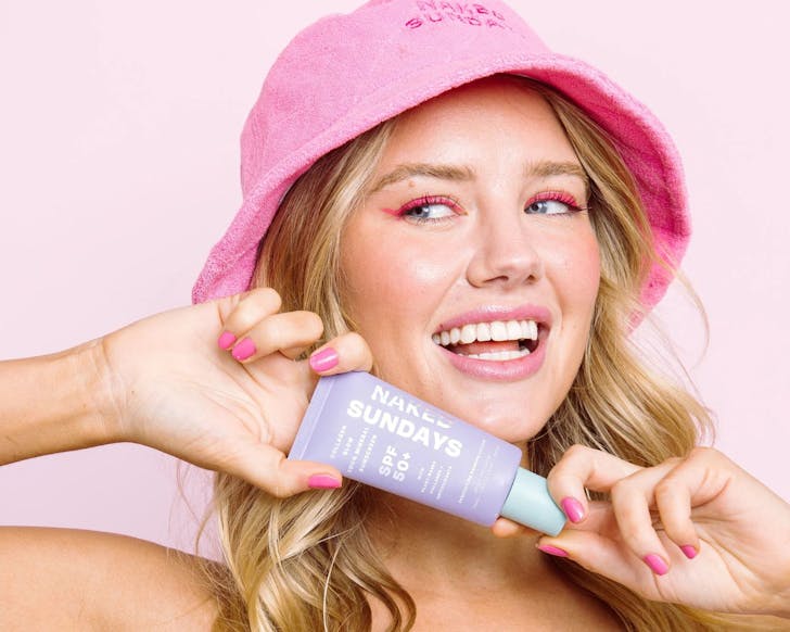 A woman in a bright hat holds a tube of Naked Sundays suncream