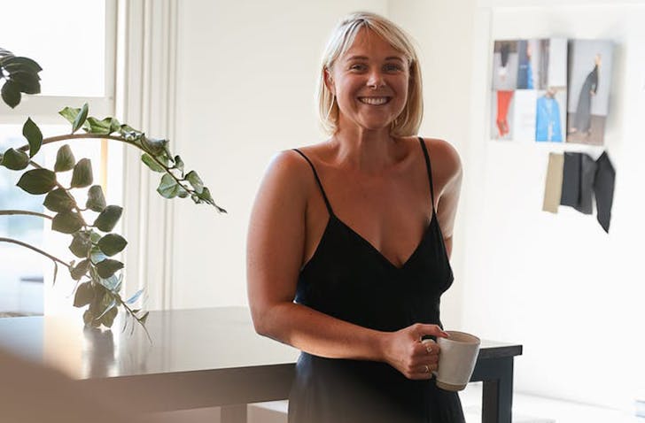 Natalie stands holding a mug of coffee, leaning on a table. Plants surround her.