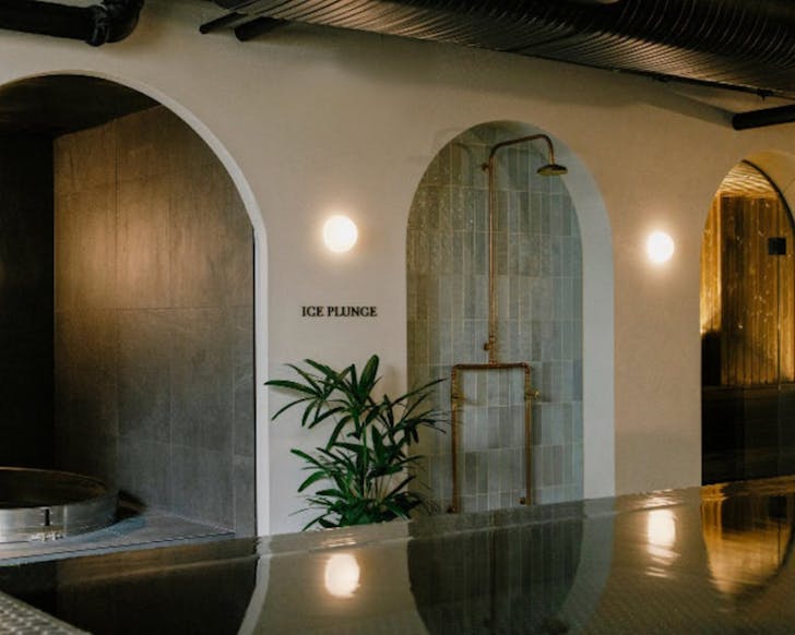 A dark bathhouse with a black tile lined spa in the forefront and cream archways at the back leading to an ice plunge and sauna
