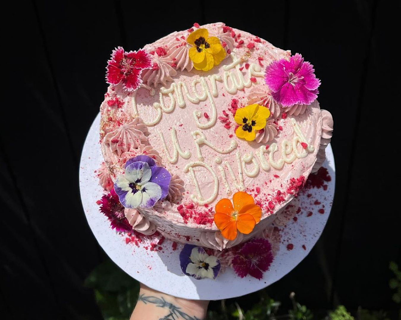 A pink cake decorated with the words “Congrats ur divorced” and fresh flowers.