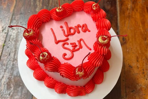A pink and red retro heart-shaped cake decorated with cherries and the words 'Libra szn.'