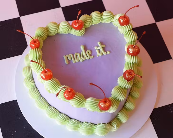 A purple vintage-style cake decorated with green icing, cherries, and the words 