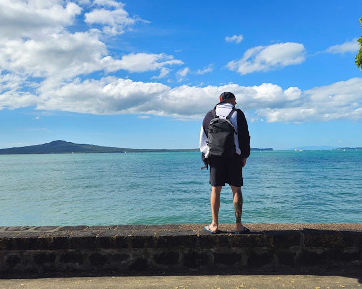 Nick stands on a rock wall at Kohimarama Beach with Rangitoto in the background.