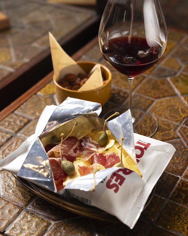 A chip packet torn open and plumped with charcuterie and olives, and a glass of red wine.