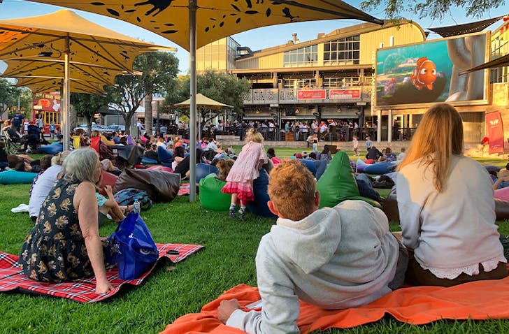 People are sitting on colourful blankets watching a movie outdoors on a projector. 