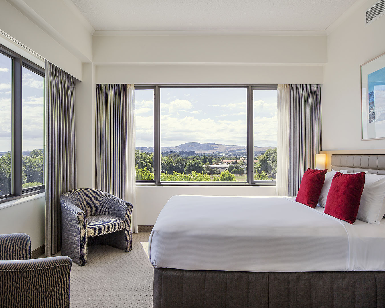 A white bed in a hotel room with great views of greenery and Lake Rotorua. 