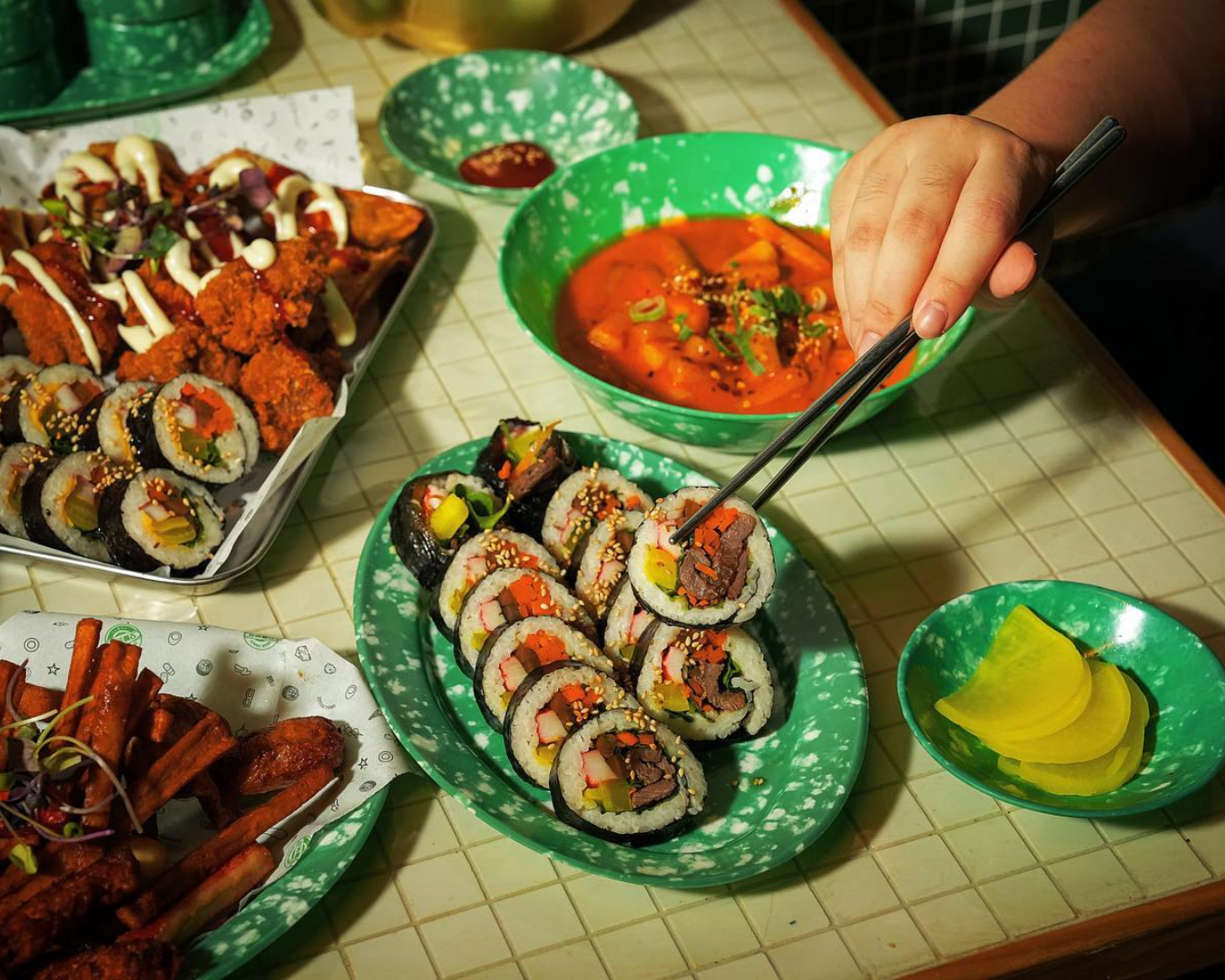 A hand holding a piece of gimbap in chopsticks with fried chicken and tteokbokki rice cakes in the background