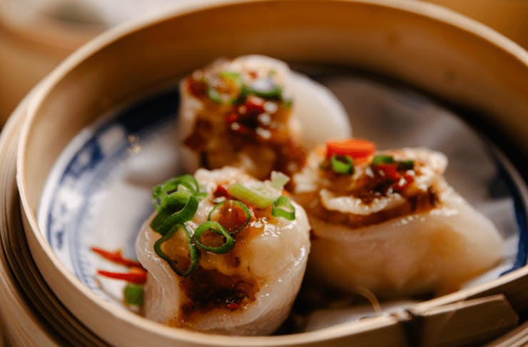 A bamboo steamer with three steamed dumplings with slices of chives and chilli resting on top. 
