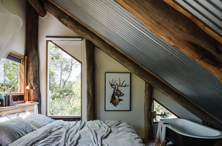 A double bed sits under a slated roof with a free-standing bath at the end.