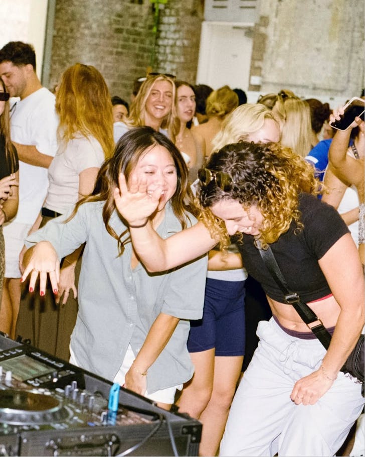 An image focusing on two girls dancing in front of a DJ desk amongst a crowd. The vibe is casual and the lighting suggests a daytime hour.