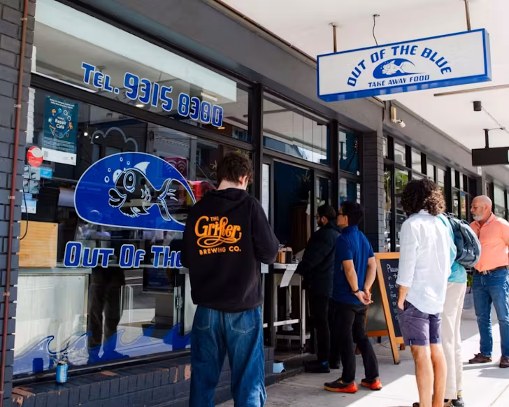 The outside of the Out Of The Blue store, with the fish logo on the front window. A line of people are outside ready to walk into the seafood shop.