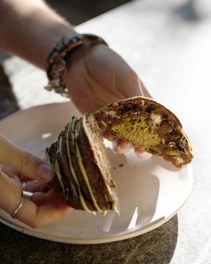 Ring adorned hands holding a chocolate coated and pistachio sauce drizzled cookie that's been cut in half, with the knafeh filled centre showing