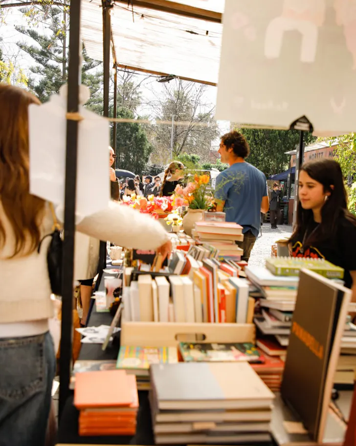 People browse a book stall at the open-air Palo Alto Market, surrounded by trees and sunlight.