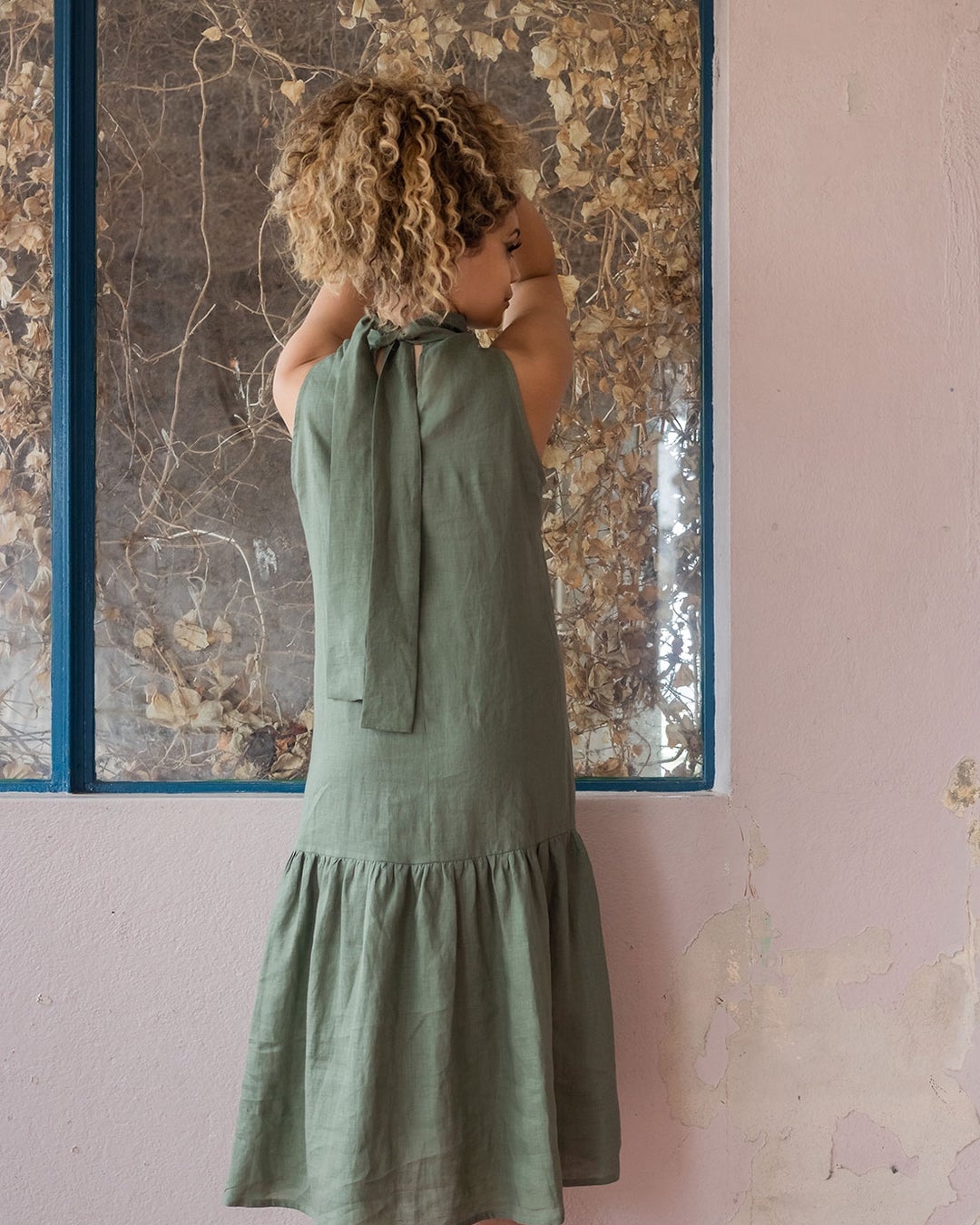 We see a woman’s back, she’s holding her hair up and looking to one side while wearing a green linen dress. 