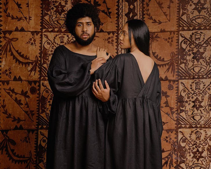 A male-presenting and woman-presenting duo stand in front of a tapa cloth background wearing black dresses by Papa Clothing. 